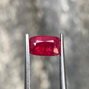Può includere: Una gemma rosso vivo, tenuta da pinzette metalliche. La gemma ha un taglio a cuscino rettangolare con sfaccettature che catturano la luce. Lo sfondo è un tono neutro sfocato, che enfatizza il colore ricco e la chiarezza della gemma.
