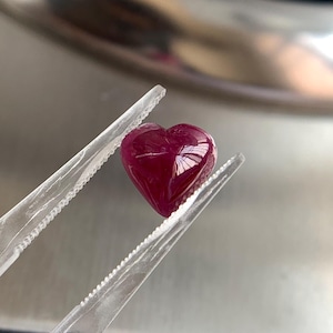 Può includere: Una gemma a forma di cuore, di colore rosso intenso, è tenuta da pinzette trasparenti. La pietra lucidata riflette la luce, mostrando la sua superficie liscia e il suo colore ricco. Le pinzette offrono una visione ravvicinata della gemma.