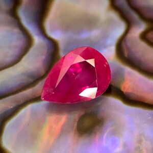 May include: A pear-shaped ruby gemstone with a deep red color. The gemstone is set against a background of iridescent abalone shell.