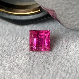 以下が含まれることがあります： 鮮やかな正方形カットのホットピンクの宝石。 宝石は、明るい灰色のテクスチャード加工された背景に配置されています。 宝石のファセットは光を反射し、その透明度と輝きを示しています。 ルビーまたはピンクサファイアである可能性があります。