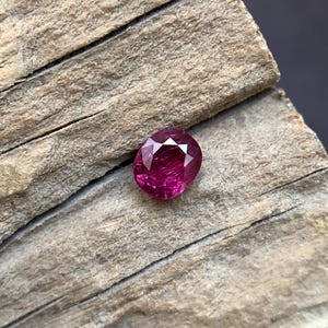 Puede incluir: Una gema facetada, de forma ovalada y color magenta intenso, descansa sobre una superficie de madera envejecida. El corte de la gema revela múltiples facetas, creando un juego de luces y sombras. La textura de la madera proporciona un contraste rústico.