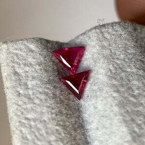 May include: Two triangular, faceted, deep red gemstones. The gems are displayed on a grey, textured fabric. The facets catch the light, creating bright highlights. The gems are likely rubies, suitable for jewellery.