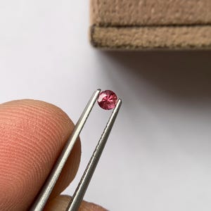 Może przedstawiać: Mały, okrągły, fasetowany, różowy kamień szlachetny trzymany jest przez srebrną pęsetę na białym tle. Kamień szlachetny ma żywy odcień różu, a fasetki tworzą efekt iskrzenia. W tle znajduje się beżowe pudełko na biżuterię.