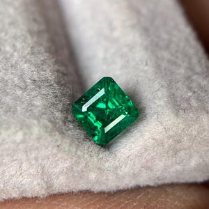 May include: A square-cut, vibrant green gemstone with faceted surfaces that reflect light. The stone displays a deep, saturated emerald hue with visible internal inclusions and sharp, clean edges, showcasing a classic geometric shape.