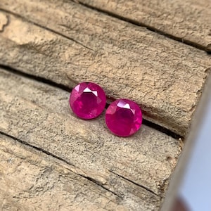 May include: Two round, faceted, bright red gemstones, possibly rubies, are shown on a rough wooden surface. The gems have a high lustre and are set against the textured, brown background.