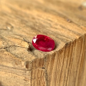 Può includere: Una gemma di rubino sfaccettata di forma ovale. La gemma è di un colore rosso intenso e vibrante, posizionata su una superficie di legno grezzo. La venatura del legno è visibile, creando un contrasto naturale con la gemma lucida.
