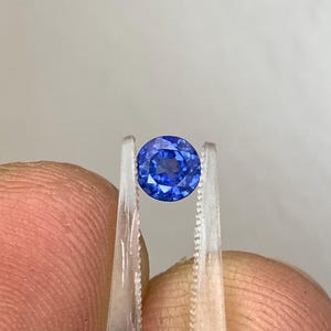 May include: A round, brilliant-cut sapphire gemstone, held by tweezers. The sapphire is a deep, vibrant blue colour, showcasing its facets. The tweezers are made of a clear material, and the background is a neutral grey.