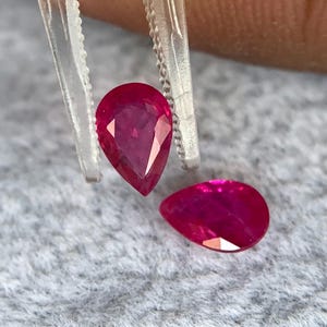 Könnte beinhalten: Zwei tropfenförmige, facettierte Rubine. Ein Rubin wird von einer transparenten Pinzette gehalten. Die Rubine haben eine tiefe, leuchtende rote Farbe. Die Edelsteine ​​werden auf einer grauen, strukturierten Oberfläche präsentiert.