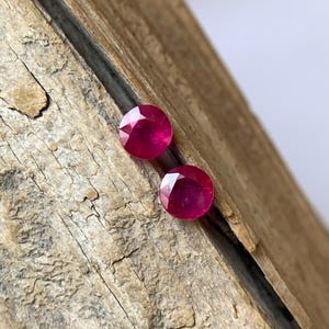 Puede incluir: Dos rubíes redondos y facetados, de color rojo intenso. Las piedras preciosas están sobre una superficie de madera texturizada de color marrón claro. Los rubíes tienen un brillo brillante, reflejando la luz.
