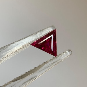 May include: A triangular, faceted, deep red gemstone held by silver tweezers. The gemstone has a sharp, geometric shape and a polished surface, reflecting light. The tweezers have a serrated edge for grip, and the background is a neutral gray.