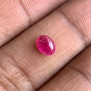May include: A vibrant, oval-shaped, ruby gemstone. The polished stone displays a deep pinkish-red hue, with internal inclusions visible. The gem is held in a hand, showcasing its size and color.
