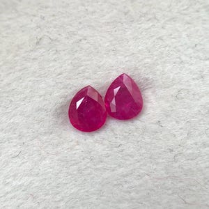 Op de afbeelding: Twee gefacetteerde, peervormige, dieprode edelstenen. De edelstenen worden gepresenteerd op een lichtgrijze, getextureerde ondergrond. De edelstenen zijn aan de onderkant puntig gesneden en hebben een afgeronde bovenkant.