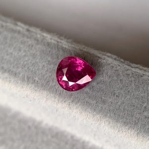 May include: A vibrant, pear-shaped ruby gemstone. The gem is a deep, saturated pink-red color, with facets that catch the light. The ruby is set against a soft, gray background, highlighting its brilliance and clarity. A close-up shot showcases the gemstone's polished surface.