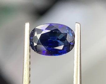 Könnte beinhalten: Ein tiefblauer ovaler Saphir-Edelstein, der in einer Pinzette gehalten wird. Der Saphir hat einen Brillantschliff und eine hohe Klarheit.