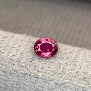 May include: A faceted, oval-cut, ruby-colored gemstone. The gem is a vibrant shade of pinkish-red, with a brilliant, reflective surface. The gemstone is set against a soft, gray background, highlighting its color and clarity. The gem is likely for jewelry.