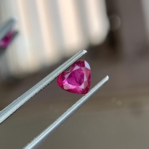 Può includere: Un rubino a forma di cuore, sfaccettato, tenuto da pinzette d'argento. Il rubino è di un vibrante colore rosso-rosato, con sfaccettature visibili che catturano la luce. Le pinzette sono a fuoco, evidenziando la forma e il colore della gemma.