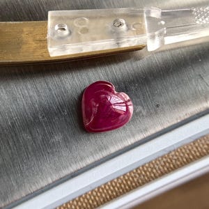Può includere: Una gemma a forma di cuore, lucidata, di colore rosso intenso. La gemma è posta su una superficie argentata, con uno strumento di plastica trasparente e uno strumento di ottone sullo sfondo. La gemma è probabilmente un rubino.