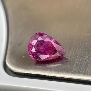 Könnte beinhalten: Ein birnenförmiger, facettierter Edelstein in einer leuchtenden, tiefrosa Farbe. Der Stein weist mehrere dreieckige und rautenförmige Facetten auf, die das Licht reflektieren und eine brillante, funkelnde Optik auf der polierten Oberfläche erzeugen.
