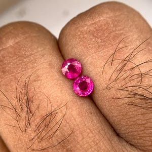 Puede incluir: Dos gemas redondas, facetadas, de color rosa brillante. Las gemas se sostienen entre los dedos, mostrando su talla brillante y su color vibrante. Las gemas parecen ser de tamaño y forma similares, reflejando la luz.