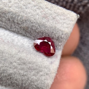 Pode incluir: Uma gema de rubi facetada em forma de coração. O rubi é de cor vermelha profunda e está sobre um tecido cinza texturizado. As facetas da gema capturam a luz, criando um efeito brilhante. Esta imagem mostra uma pedra preciosa.