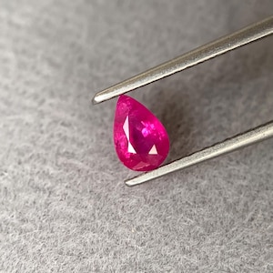 May include: A vibrant, pear-shaped ruby gemstone, held by silver tweezers. The ruby displays a deep, saturated red hue, with facets catching the light. The background is a soft, textured gray surface.