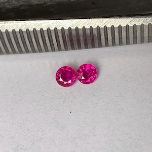 May include: Two round, vibrant pink gemstones are displayed on a white surface. The gems are positioned near a metal tool with a ridged edge, likely used for handling or setting the stones. The gems are faceted, reflecting light.
