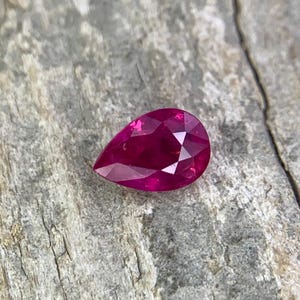May include: A pear-shaped ruby gemstone with a deep red colour. The gemstone is set against a rough, textured background.