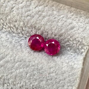 May include: Two round, faceted, deep red gemstones, possibly rubies, are presented against a soft, grey, textured backdrop. The gems are placed close together, highlighting their brilliant cut and rich colour. The setting appears to be a jewellery box.