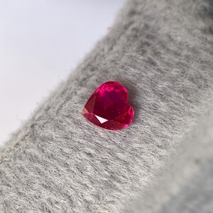 May include: A heart-shaped, faceted ruby gemstone. The ruby is a deep, vibrant red color and sits on a gray, textured surface. The gemstone's facets catch the light, creating a sparkling effect. The heart shape is clearly defined.