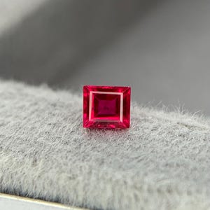 Puede incluir: Una gema roja vibrante de corte cuadrado. La gema tiene un color profundo y rico y una forma precisa y simétrica. La piedra preciosa se encuentra sobre un fondo gris suave, resaltando su brillo.