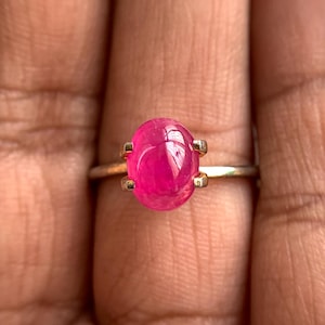 May include: A gold ring featuring a large, oval-shaped, vibrant pink gemstone. The gemstone is held in place by four gold prongs, and the band is a simple, thin gold. The ring is shown on a person's finger.