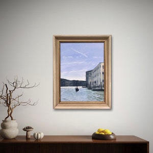 May include: A framed painting of a canal in Venice, Italy. The painting depicts a gondola in the water with buildings on either side of the canal. The sky is blue with a few white clouds.