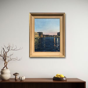 May include: A framed painting of a canal in Venice, Italy. The painting depicts a cityscape with buildings lining the canal, and the water is reflecting the light from the setting sun.