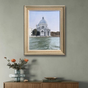 May include: A framed painting of a white building with a dome, situated on a body of water. The building is surrounded by trees and other buildings. The water is a light blue-green color and reflects the sky.