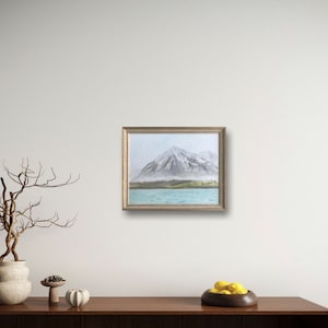 May include: A framed painting of a snow-capped mountain with a lake in the foreground. The mountain is a light gray color with a hint of green at the base. The lake is a light blue color and the sky is a light gray color.