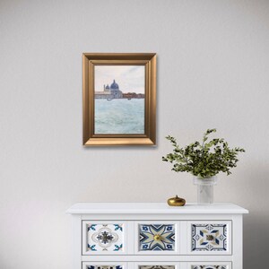 May include: A framed painting of a cityscape with a large building with a dome in the background. The painting is in a gold frame and is hanging on a white wall.