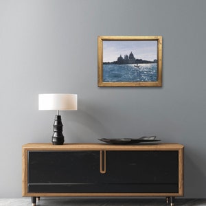May include: A framed painting of a cityscape with a boat in the water. The painting is in a gold frame and is hanging on a gray wall. A lamp and a black and wood cabinet are in the foreground.