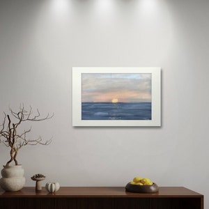 May include: A framed painting of a seascape with a setting sun over the ocean. The sky is a soft blue and pink, and the water is a deep blue. The sun is a bright yellow.