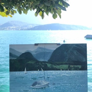 May include: A postcard with a painting of a lake with mountains in the background and sailboats on the water. The postcard is propped up on a wooden surface with a view of a lake and mountains in the distance.