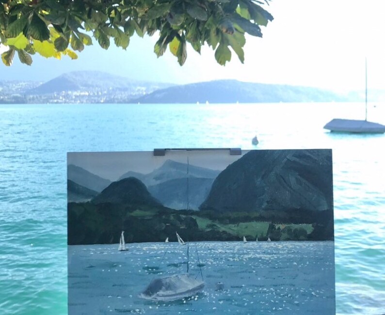 May include: A postcard with a painting of a lake with mountains in the background and sailboats on the water. The postcard is propped up on a wooden surface with a view of a lake and mountains in the distance.