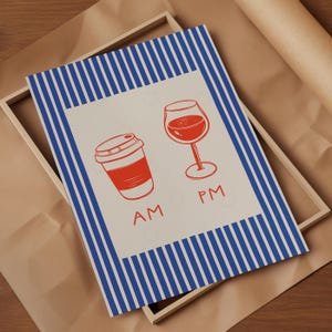 May include: A framed print with a blue and white striped border. The print features a red coffee cup and a wine glass, with "AM" and "PM" written below. The background is a light beige color.