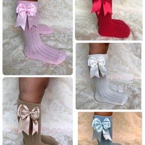 Könnte beinhalten: Eine Auswahl an kniehohen Socken in verschiedenen Farben, jeweils mit einer großen Satinschleife am Bund. Farben sind Rosa, Rot, Weiß, Beige und Blau. Die Socken haben eine gerippte Textur.