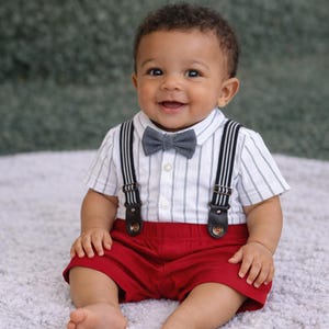 Könnte beinhalten: Ein lächelndes Baby in einem Outfit mit einem weiß-schwarz gestreiften Hemd, einer grauen Fliege und schwarzen Hosenträgern. Das Baby trägt auch rote Shorts. Ein klassischer, stilvoller Look für einen besonderen Anlass.