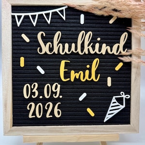 Könnte beinhalten: Ein quadratisches schwarzes Filz-Letterboard mit hellem Holzrahmen, verziert mit weißen und gelben Ausschnitten wie Wimpelketten, Konfetti und einer Schultüte. Der Text lautet Schulkind, Emil, 03.09.2026 in weißen und gelben Schrift- und Blockbuchstaben.