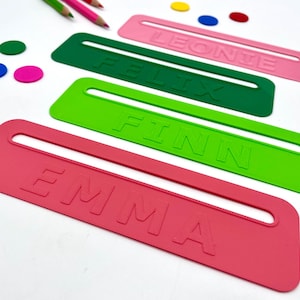 Könnte beinhalten: Vier farbige Namensschilder in Pink, Grün und Limettengrün, jedes mit einem anderen Namen geprägt: Emma, Finn, Felix und Leonte. Die Schilder haben eine rechteckige Form mit einem Ausschnitt für eine einfache Handhabung. Stifte und farbige kreisförmige Akzente sind verstreut.