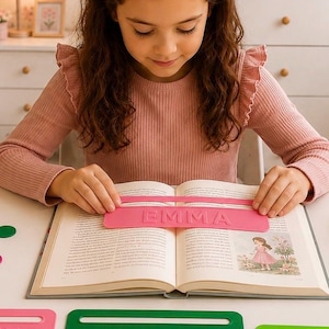 Könnte beinhalten: Eine rechteckige Lesehilfe aus rosa Kunststoff mit dem eingeprägten Namen EMMA in Großbuchstaben, die horizontal auf einem aufgeschlagenen Buch liegt. Weitere rechteckige Lesehilfen in Rosa und Grün mit Namen wie FELIX sind zu sehen.