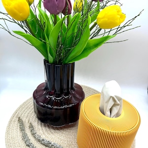Könnte beinhalten: Ein Stillleben mit einer dunkelvioletten Vase mit gelben und lila Tulpen und grünen Blättern. Daneben eine gelbe, gerippte Taschentuchbox. Die Anordnung befindet sich auf einem beigen Platzset.
