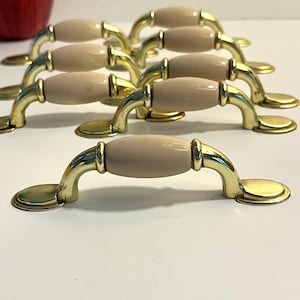 May include: A collection of eight cabinet pulls. Each pull features a gold-toned metal handle with a light beige, oval-shaped center. The pulls are arranged in rows, showcasing their design and finish. The pulls are designed for furniture.