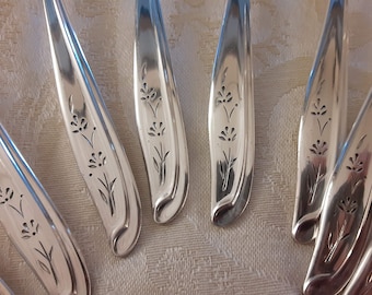 May include: Close-up of six silver spoons with floral designs etched into the handles. The spoons are arranged in a fan shape on a white tablecloth.