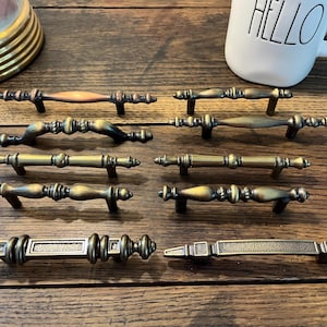 May include: A collection of antique-style cabinet pulls in various designs and finishes. The pulls are made of metal and feature ornate details, with colors ranging from bronze to gold. The pulls are arranged on a wooden surface.